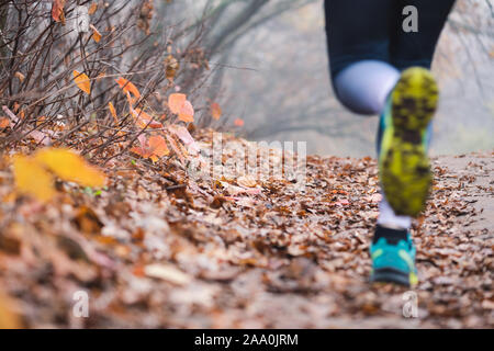 Female jogger legs feets close up back. Running woman legs in defocus, Fallen foliage on the ground, autumn foggy morning. Shooting point from below. Stock Photo