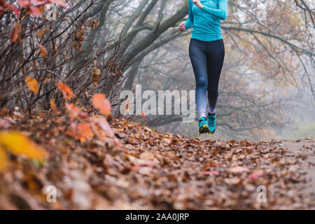 Woman jogging jumping in forest, no face. Fallen foliage on the ground, autumn foggy morning. Shooting point from below. Flying runner. Stock Photo