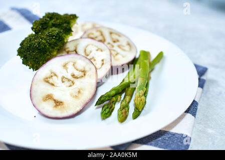 Steamed vegetables in a white plate on a blue table. Eggplant, broccoli, asparagus. Stock Photo