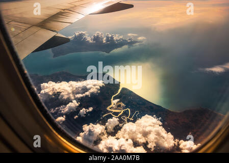 Aerial View from Window of Airplane. Flying above Clouds and Beautiful Land with River Delta and Sea at Sunrise. Stock Photo