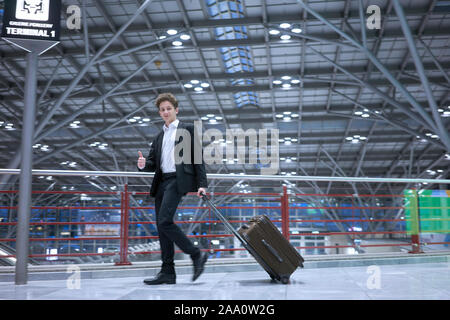 Young handsome businessman with suit walking happily through airport with rolling suitcase Stock Photo