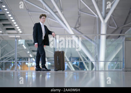 Young relaxed smiling businessman standing with his rolling suitcase in the airport, leaning with his arm on a guard railing Stock Photo