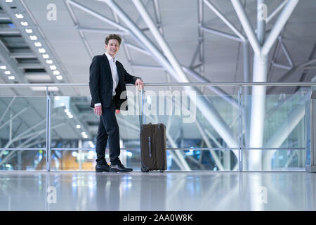 Young relaxed smiling businessman standing with his rolling suitcase in the airport, leaning with his arm on a guard railing Stock Photo