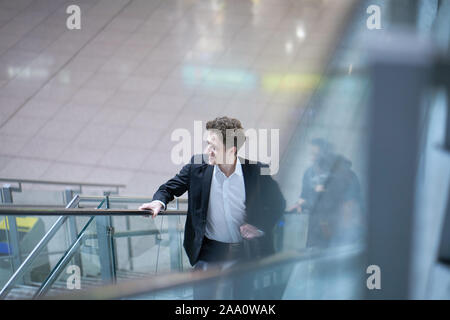 Young handsome smiling businessman inside the airport on an escalator with his rolling suitcase Stock Photo