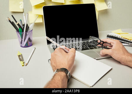cropped view of man holding glasses while writing on notebook near laptop with blank screen and stationery on grey and white Stock Photo