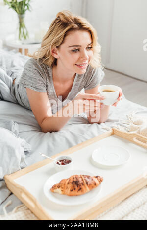 selective focus of cheerful girl holding cup of coffee near tray with tasty croissant on bed Stock Photo