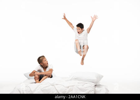 excited boy levitating over shocked brother sitting on bedding isolated on white Stock Photo