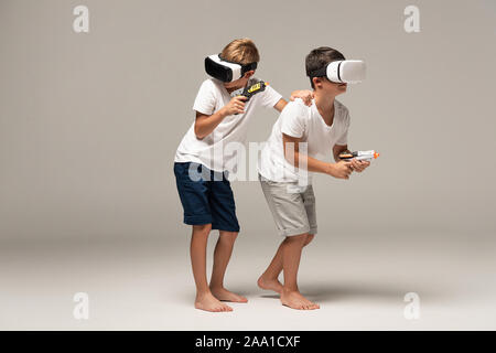 two barefoot brothers in pajamas holding toy guns while using vr headsets on grey background Stock Photo
