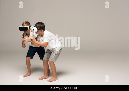 two barefoot brothers in pajamas slinking with toy guns while using vr headsets on grey background Stock Photo