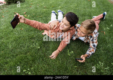 cheerful boy taking selfie on smartphone while lying on grass with brother Stock Photo