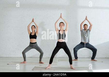 young men and woman practicing yoga in goddess pose with raised prayer hands Stock Photo