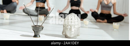 selective focus of decorative buddha head, aromatic sticks and candles, and young people practicing yoga in half lotus pose, panoramic shot Stock Photo