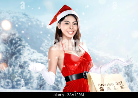 Asian woman in Santa costume holding shopping bags with Boxing Day Sale text with snowy hill background Stock Photo