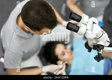 Smart dentist in a gray uniform with white latex gloves treats his female patient in a blue bib in a clinic. There is a dental microscope next to him. Stock Photo