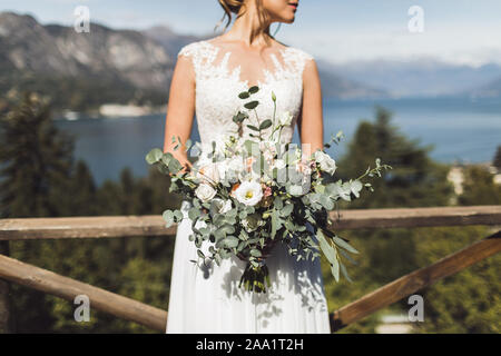Woman holding in hands big wedding bouquet in rustic style. Greens and white roses. Lake and mountain view on background. Stock Photo