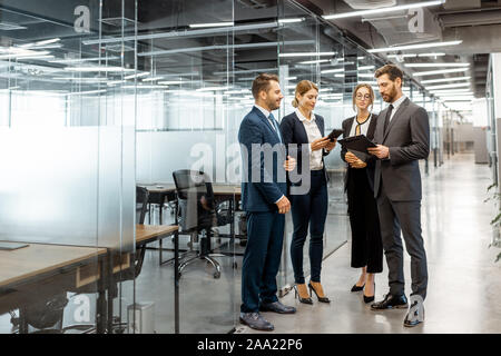 Group of business people strictly dressed in the suits meeting in the hallway of the modern office building, white-collar workers having informal discussion indoors Stock Photo