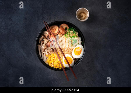 Ramen. Soba noodles with eggs, shiitake mushrooms, and vegetables, overhead shot on a black background with chopsticks and a cup of sake Stock Photo