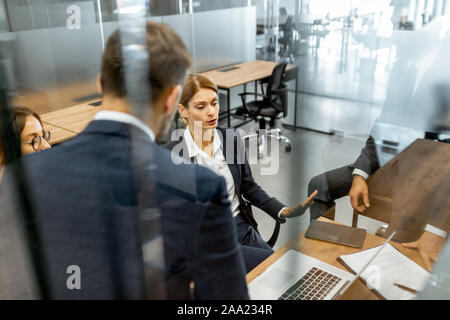 Group of strictly dressed business people having a discussion, sitting at the workplace with a computer in the modern office with glass partitions Stock Photo
