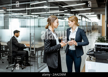 Two business ladies talking about emplyoees near the copier during a coffee break in the hallway of the big corporation Stock Photo