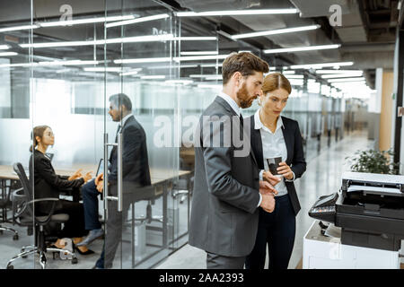 Business man and woman talking near the copier during a coffee break in the hallway of the big corporation Stock Photo