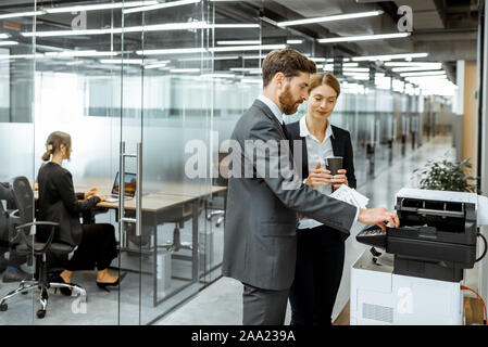 Business man and woman talking near the copier during a coffee break in the hallway of the big corporation Stock Photo