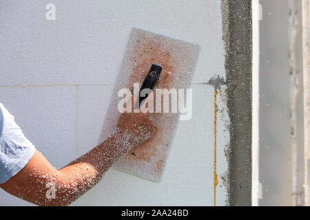 Hand of a worker with a tool for sanding and making smooth and even the surface of a house wall insulated with styrofoam insulation sheets. Stock Photo