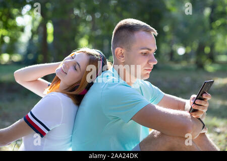 Young teenage couple having fun outdoors in summer park. Girl with red hair listening to music in pink earphones and boy chatting on the sell phone. Stock Photo