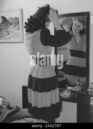 Women's fashion in the 1940s. A young dark haired woman in front of the mirror fixes her hair and uses the fashionable hair net of the time. The dress is long armed with wide horizontal stripes of dark and lighter fabric. Sweden 1945 Stock Photo