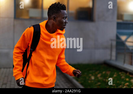 african-american man in stylish orange hoodie sweatshirt with backpack on city street in the evening Stock Photo