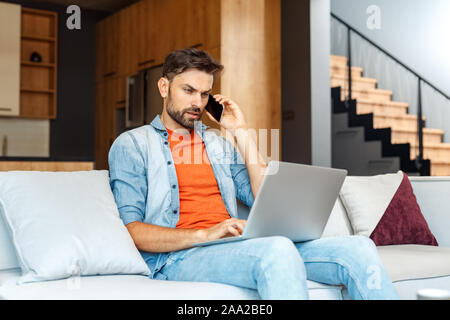 Young adult man talking on smartphone, using laptop, working at home Stock Photo