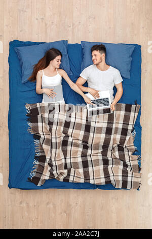Bedtime. Young man watching movie on laptop while woman with book pointing at screen smiling cheerful lying on bed under blanket top view Stock Photo