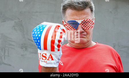 Brutal, proud, caucasian ethnicity senior man in glasses in the colors of American flag. He threatens someone with his fist in boxing glove. Close-up. Stock Photo