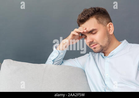 Freestyle. Young man studio sitting on sofa isolated on grey touching head having headache tired close-up Stock Photo