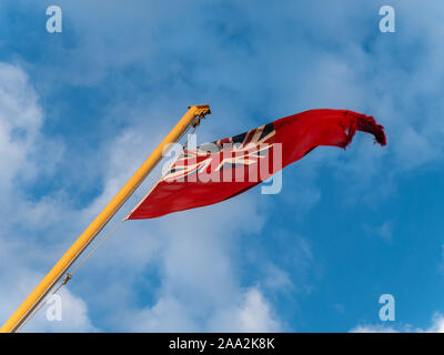 Frayed British Red Ensign flag flying from flagstaff against blue sky. Stock Photo