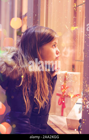 Beautiful little girl in front of confectionery shop, decorated with wrapped gift boxes and candies. Christmas concept, window shopping Stock Photo