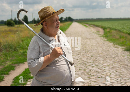 Nice outdoor portrait of Ukrainian senior farmer in straw hat standing on cobblestone road and taking walking stick Stock Photo