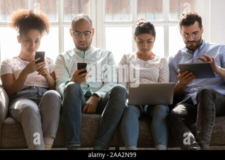 Concentrated diverse people sitting together on sofa, using different gadgets. Stock Photo