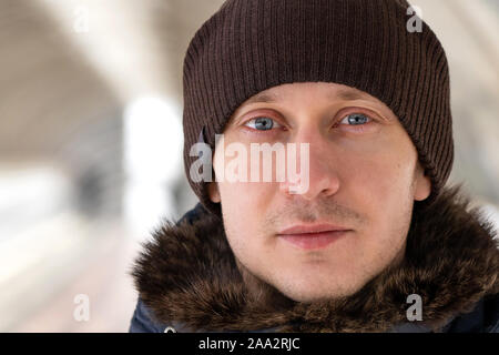 Close up street portrait of a young unshaven European man with gray eyes wearing a knitted warm hat and warm winter jacket, standing in the transition Stock Photo