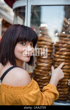 Beautiful young girl in fashionable clothes shows stall of traditional Turkish street food simit or bagel in English at a steet.