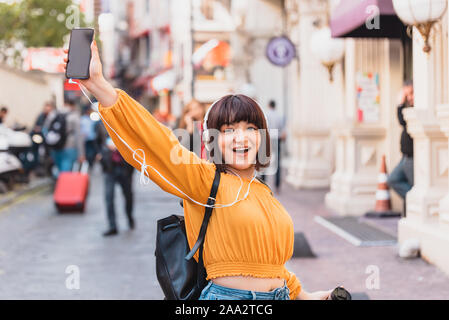 Beautiful young girl in fashionable clothes listen music with coffee mug while standind at street Stock Photo