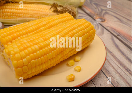 Two boiled ears of corn on a plate and raw ears of corn on a wooden table. Healthy diet. Fitness diet. For a sweet treat. Close up Stock Photo