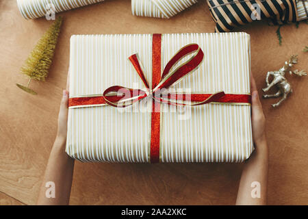 Hands giving stylish christmas gift box in striped golden paper  with red bow, and presents, golden tree, deer on wooden table. Wrapping christmas gif Stock Photo