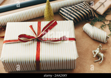 Stylish christmas gift box in striped golden paper and with red bow, and scissors, presents, golden tree, deer on wooden table. Wrapping christmas gif Stock Photo