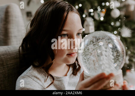 Girl sitting by a Christmas tree looking at a snow globe Stock Photo