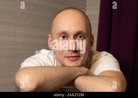 A 30-year-old man without hair laid his chin in his hands and looks at the camera. Portrait of a young man in a white t-shirt. Stock Photo