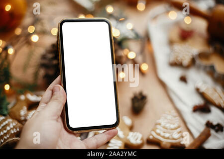 Phone with empty screen in hand on background of christmas lights, festive gingerbread cookies, anise, cinnamon, pine cones, cedar branches on rustic Stock Photo