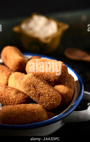 closeup of some spanish croquettes in a white and blue enamel plate placed on a rustic wooden table, and a small yellow mortar with aioli sauce in the Stock Photo
