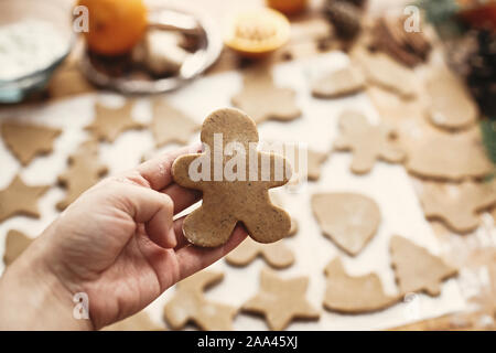 Hand holding raw gingerbread man cookie on background of dough, metal cutters and anise, ginger, cinnamon, pine cones, fir branches on rustic table.Ma Stock Photo