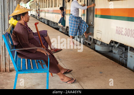 An elderly Burmese woman dressed in a traditional conical Asian hat and holding a cane waits to board a train at the Yangon Central Railroad station Stock Photo