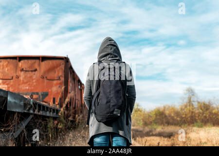 Homeless woman with backpack getting away from it all, rear view of female walking among abandoned train wagons and obsolete railroad track Stock Photo